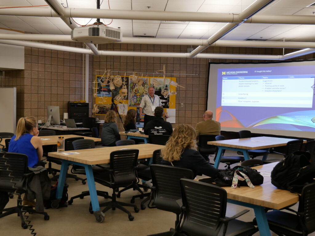A speaker presents a slideshow to an audience in a classroom with movable tables.